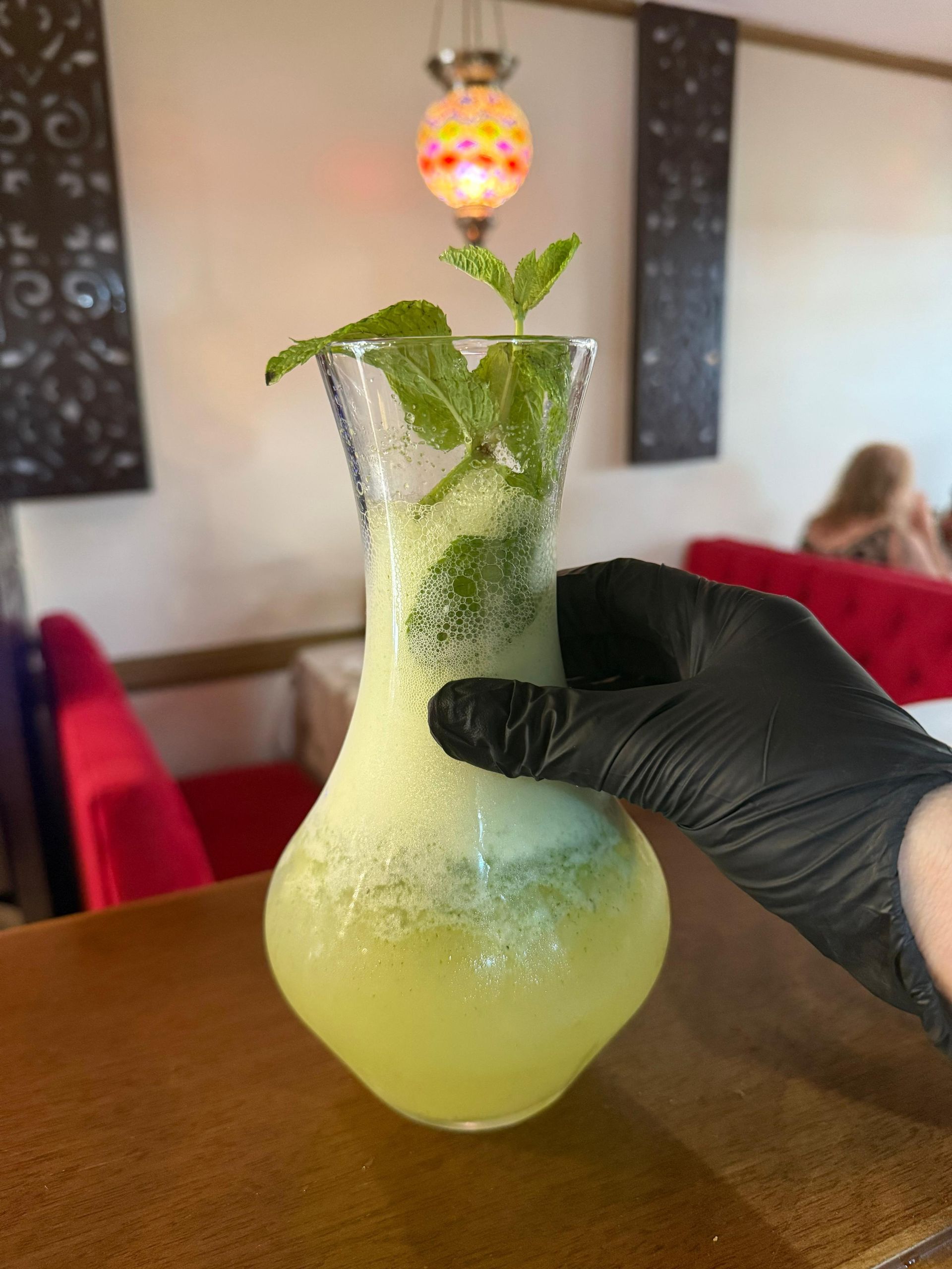Cóctel verde en un vaso helado con menta y una guarnición colgante, sostenido por una mano enguantada.