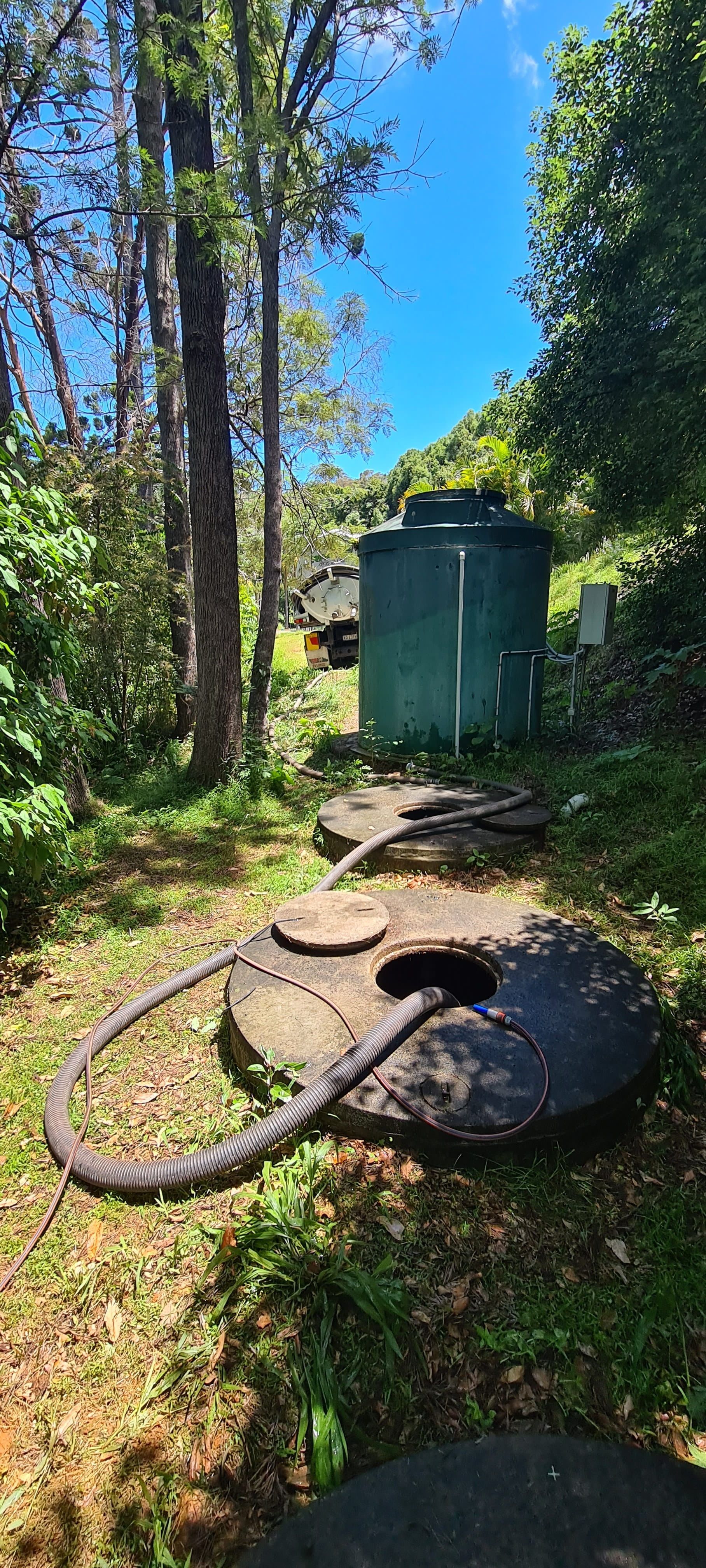 A grassy outdoor scene with a dark green water tank, open septic tank lids, and tall trees under a blue sky — Raptor Waste Management in Coolangatta, QLD