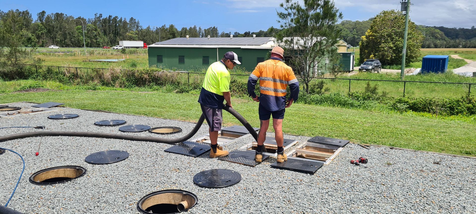 Two Workers, One Holding a Hose, Servicing Septic Tanks in a Gravel Area Near a Green Building — Raptor Waste Management in Coolangatta, QLD