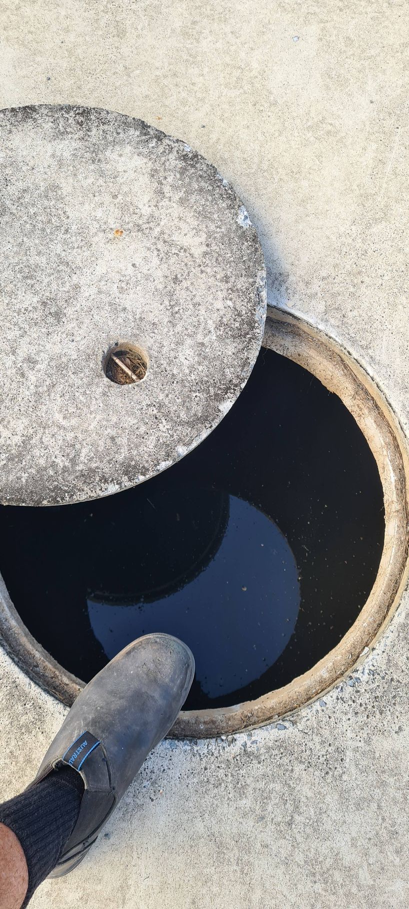 A man's foot wearing a gray boot over an open manhole with dark liquid reflecting a crescent shape — Raptor Waste Management in Coolangatta, QLD