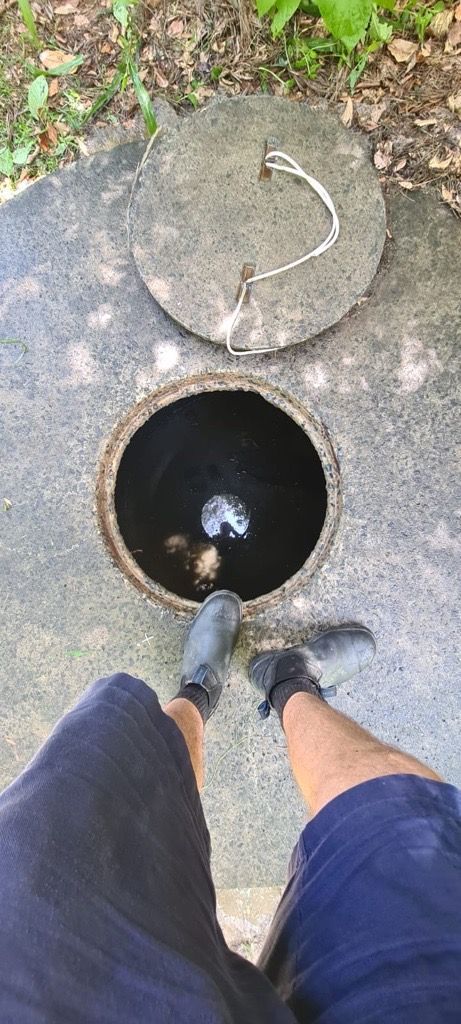 Person standing over an open manhole, with the lid ajar. Concrete ground, dark hole, blue shorts. — Raptor Waste Management in Coolangatta, QLD