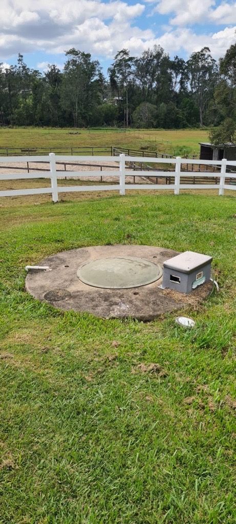 Concrete septic tank lid in a grassy yard, with a small utility box beside it, white fence and trees in background. — Raptor Waste Management in Coolangatta, QLD