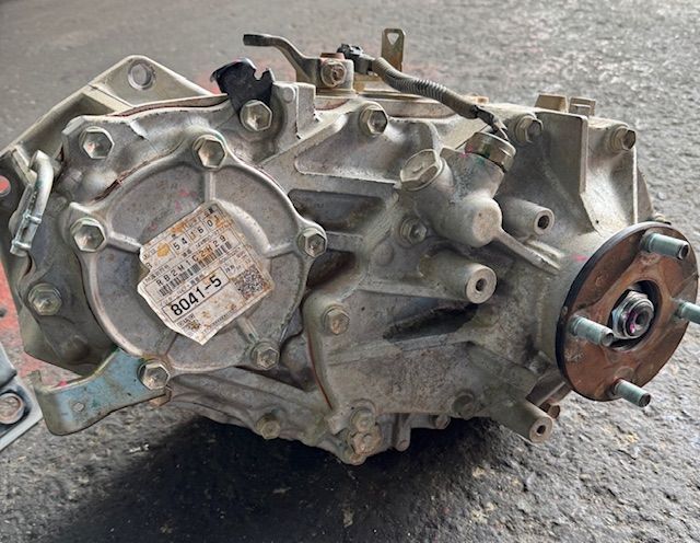 Transfer case for a vehicle, with visible bolts, metal casing, and attached components. A sticker is on the main face of the case.