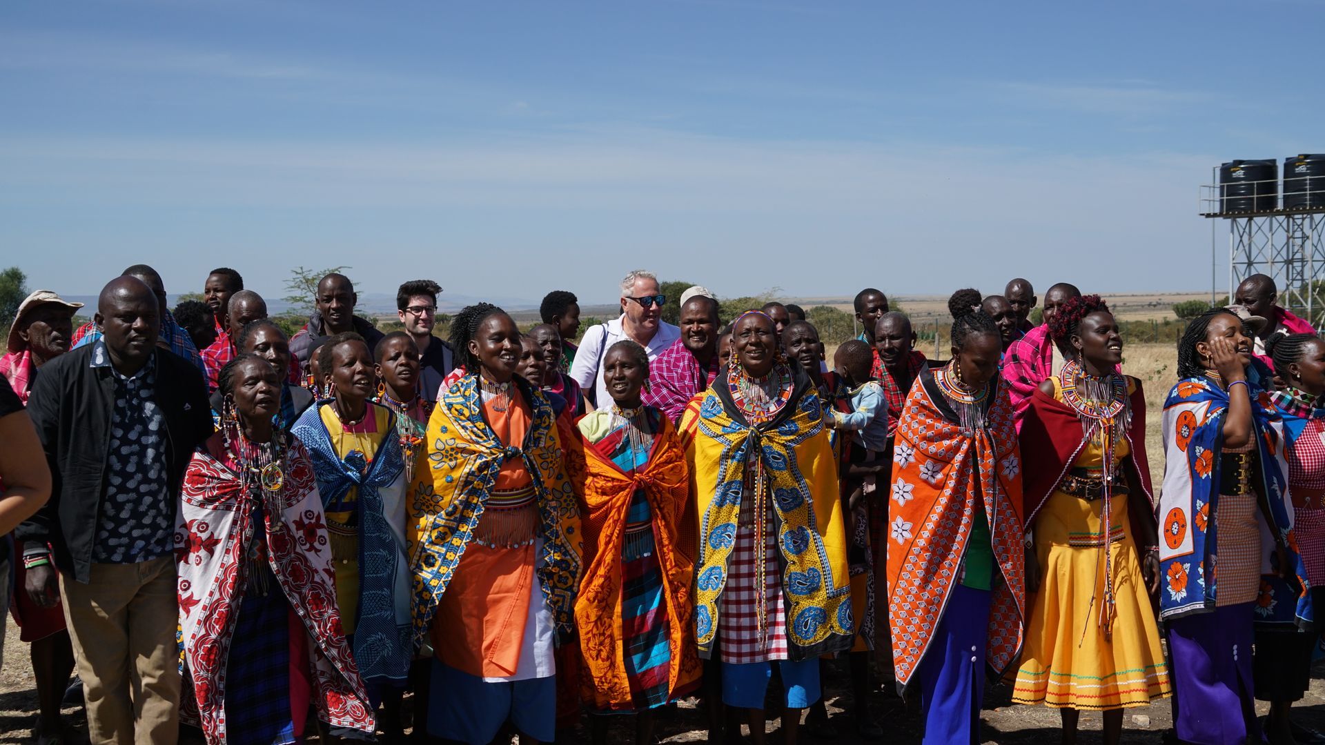 A large group of people are standing next to each other in a field.  A group of children in school uniforms are holding bouquets of yellow flowers.  Andy Stein with Orphaned Starfish Foundation (OSF) helping children in the African Continent including Kenya,  Masai Mara Tribe, Tanzania, and Ethiopia.