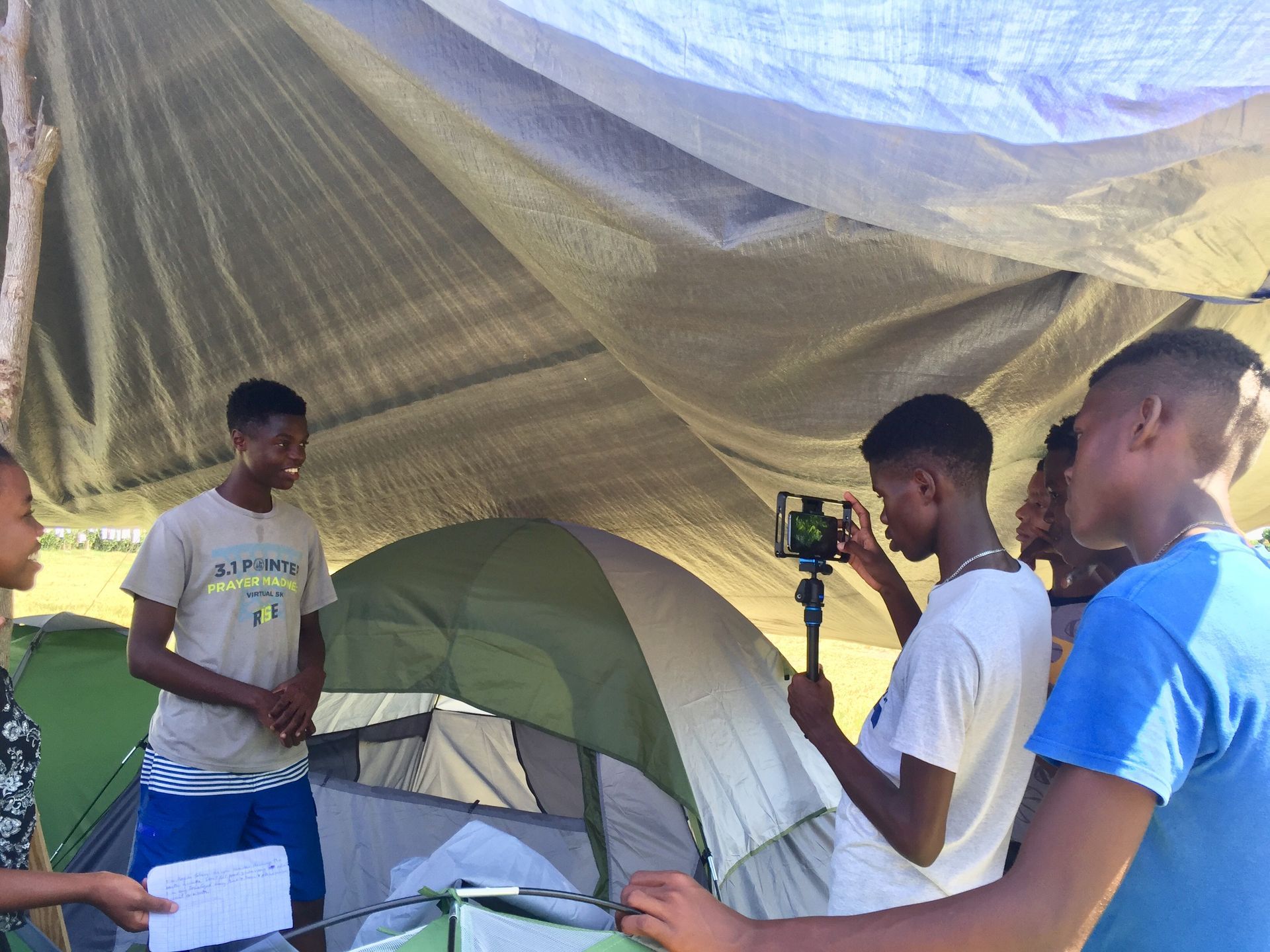 A group of young men are standing under a tarp in front of a tent creating and producing a movie.