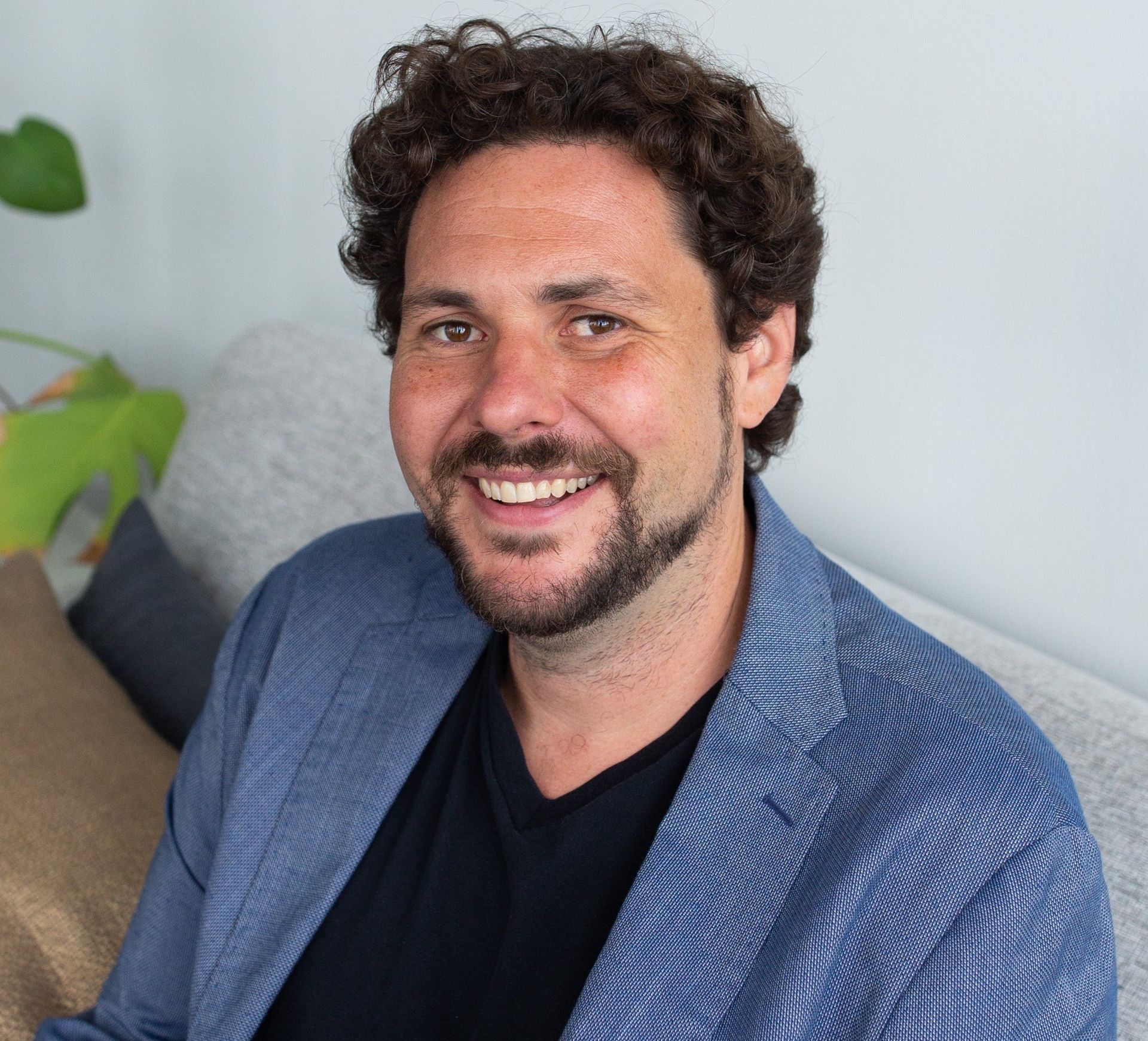 A man with curly hair and a beard is smiling while sitting on a couch.