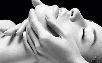 Black and white image of womans face with a spa technicans hands on her face, give her a facial at a beauty spa