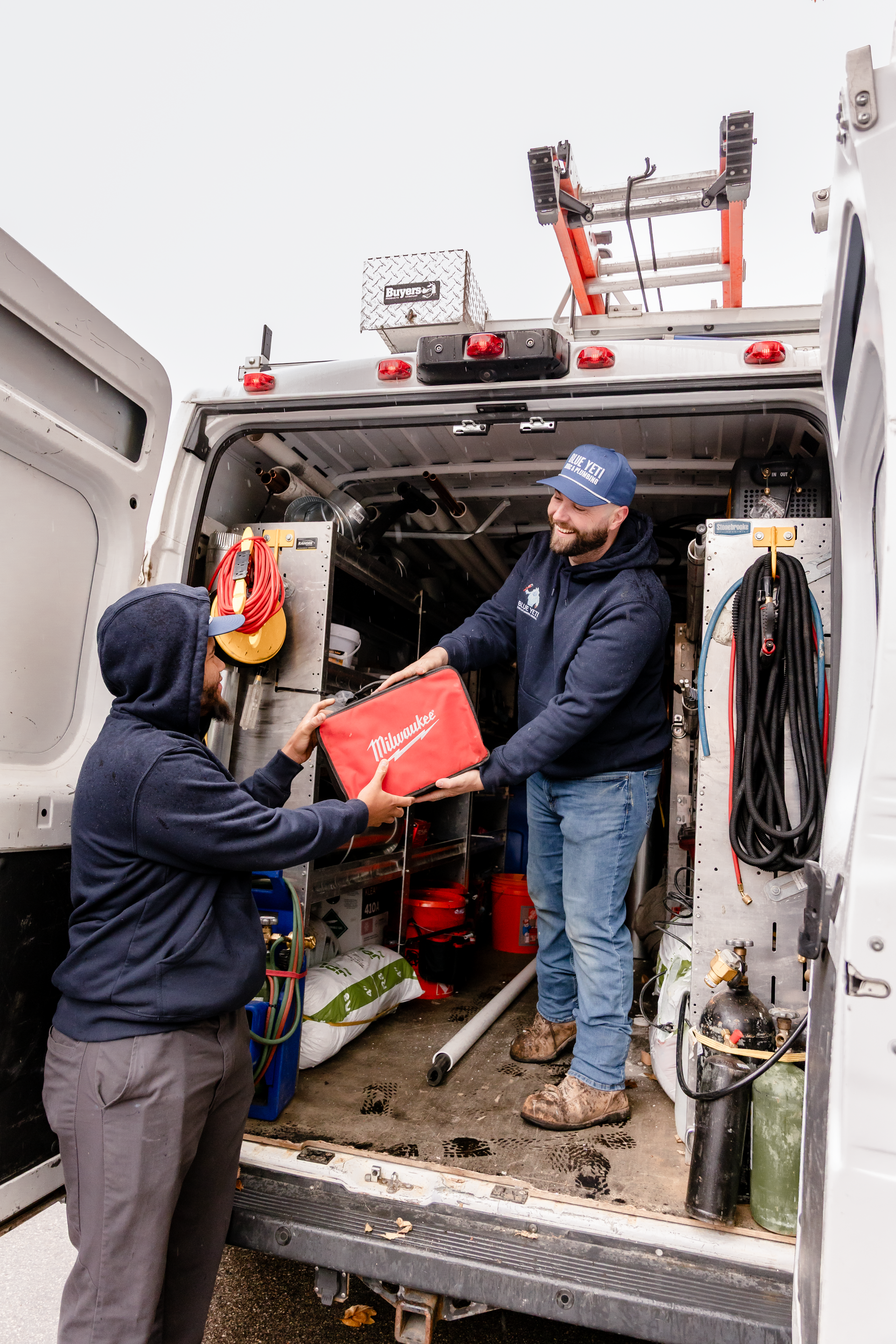 Two Blue Yeti HVAC Plumbing HVAC technicians handing each other a red tool case for a service job