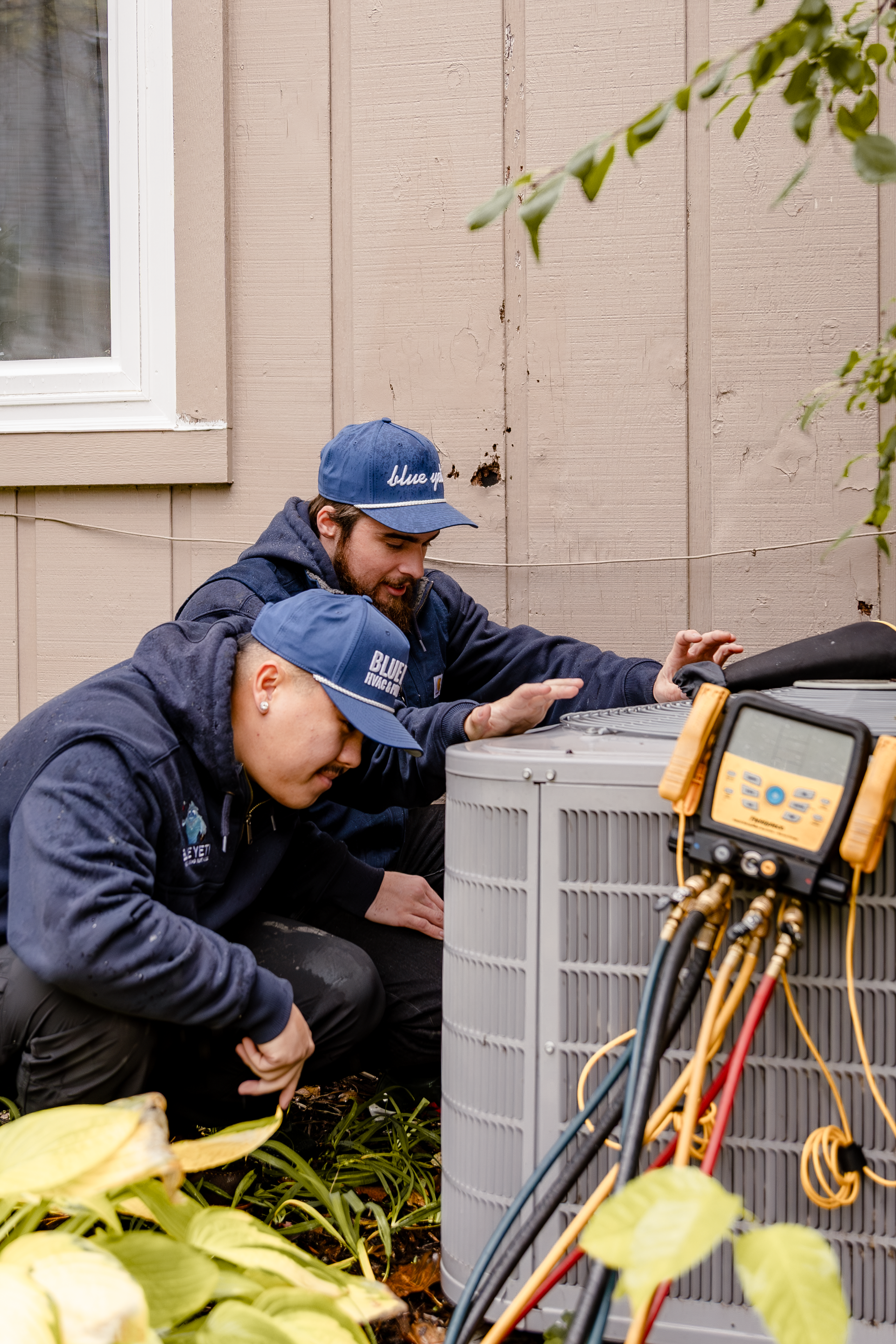 Two Blue Yeti HVAC Plumbing technicians in blue hats and jackets inspect an AC unit outside a building
