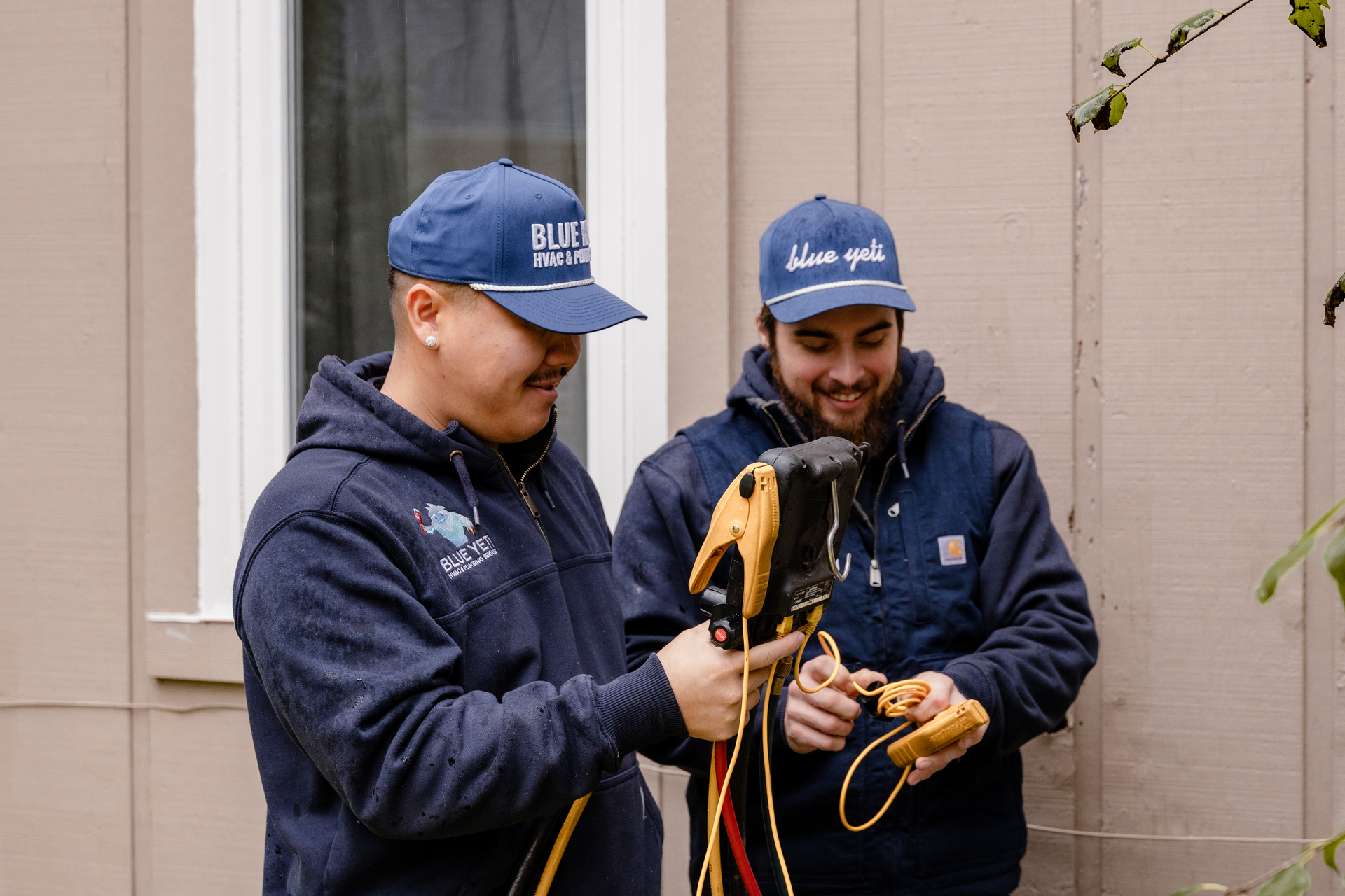 Two Blue Yeti HVAC Plumbing HVAC technicians in blue hats and jackets examining equipment outdoors