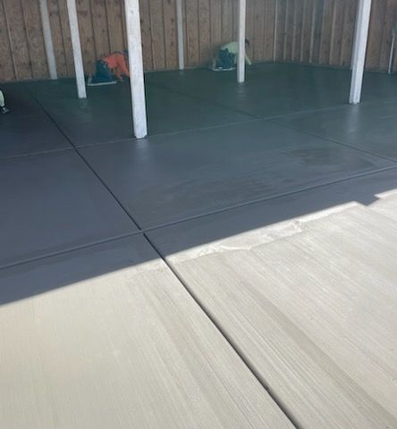 Freshly poured concrete patio with dark grey and light grey sections, divided by lines, under wooden fence and white support beams.