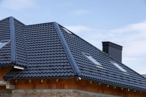 Dark gray tiled house roof with dormer windows and chimney under a blue sky