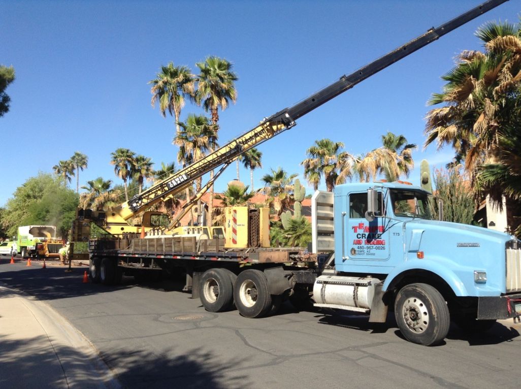 Light blue flatbed truck with crane on a street, hauling wooden boxes. Palm trees in the background.