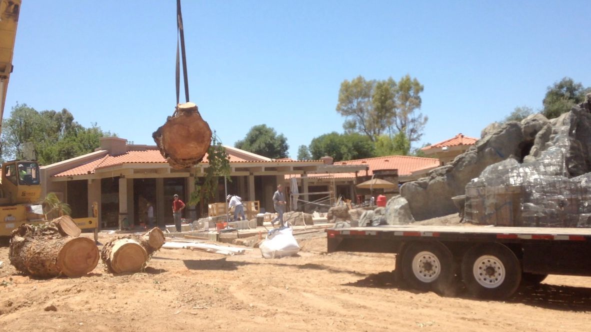 A crane lifting a large, dirt-covered object over a construction site with workers and a house under renovation.
