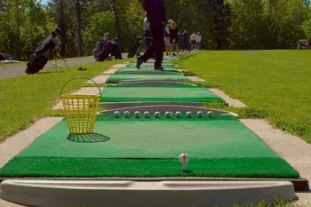 Golfer swinging at a golf ball on a driving range, with other golfers and trees in the background.