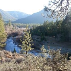 River winds through a valley surrounded by mountains and evergreen trees. Sunny day, light haze.