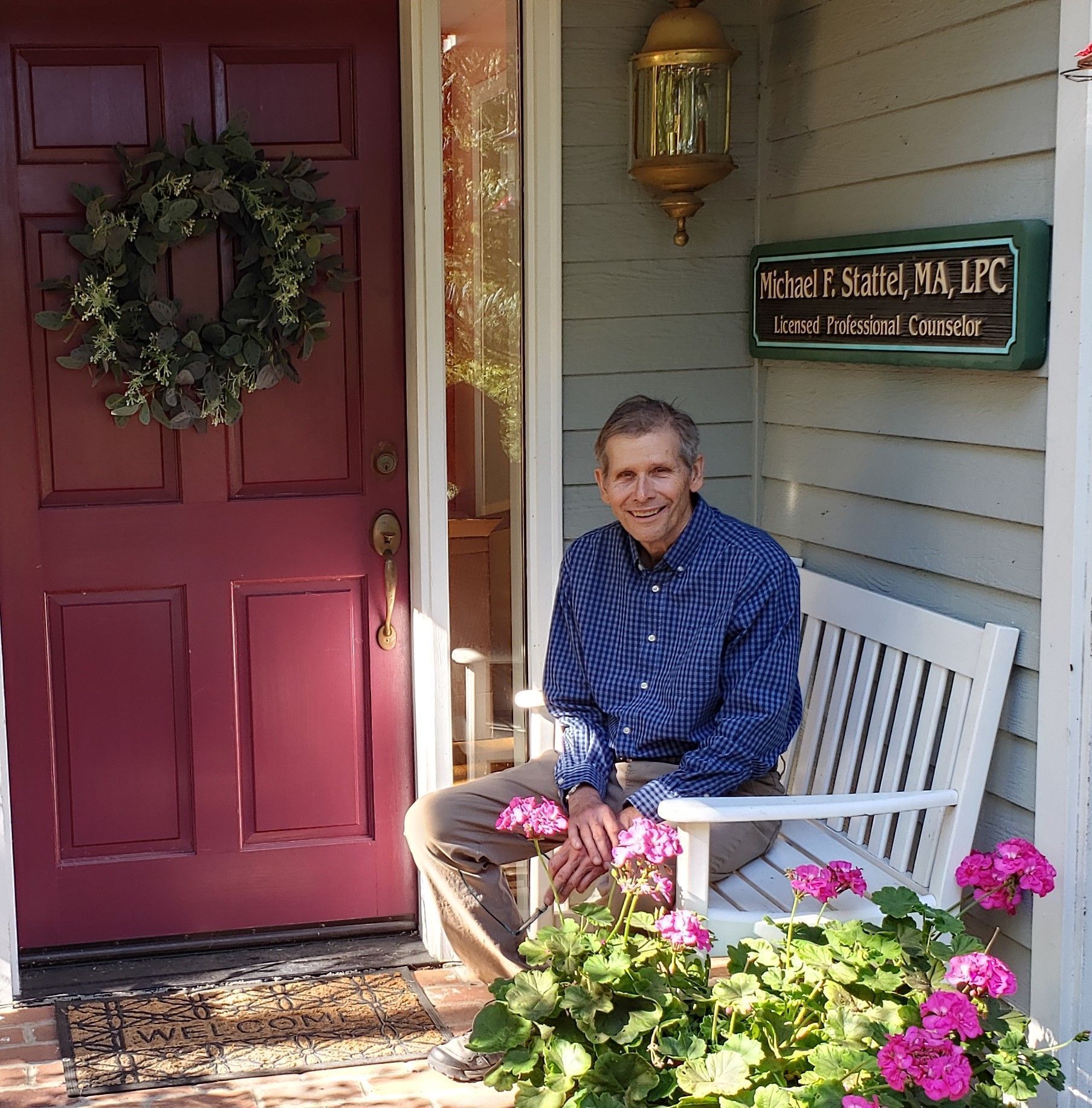 Man sitting on a white bench on a porch with a wreath, red door, and sign for a therapist.