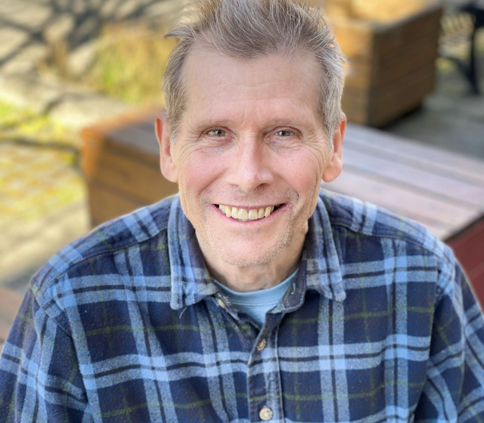 Man with graying hair and blue plaid shirt smiles outdoors, blurry background.
