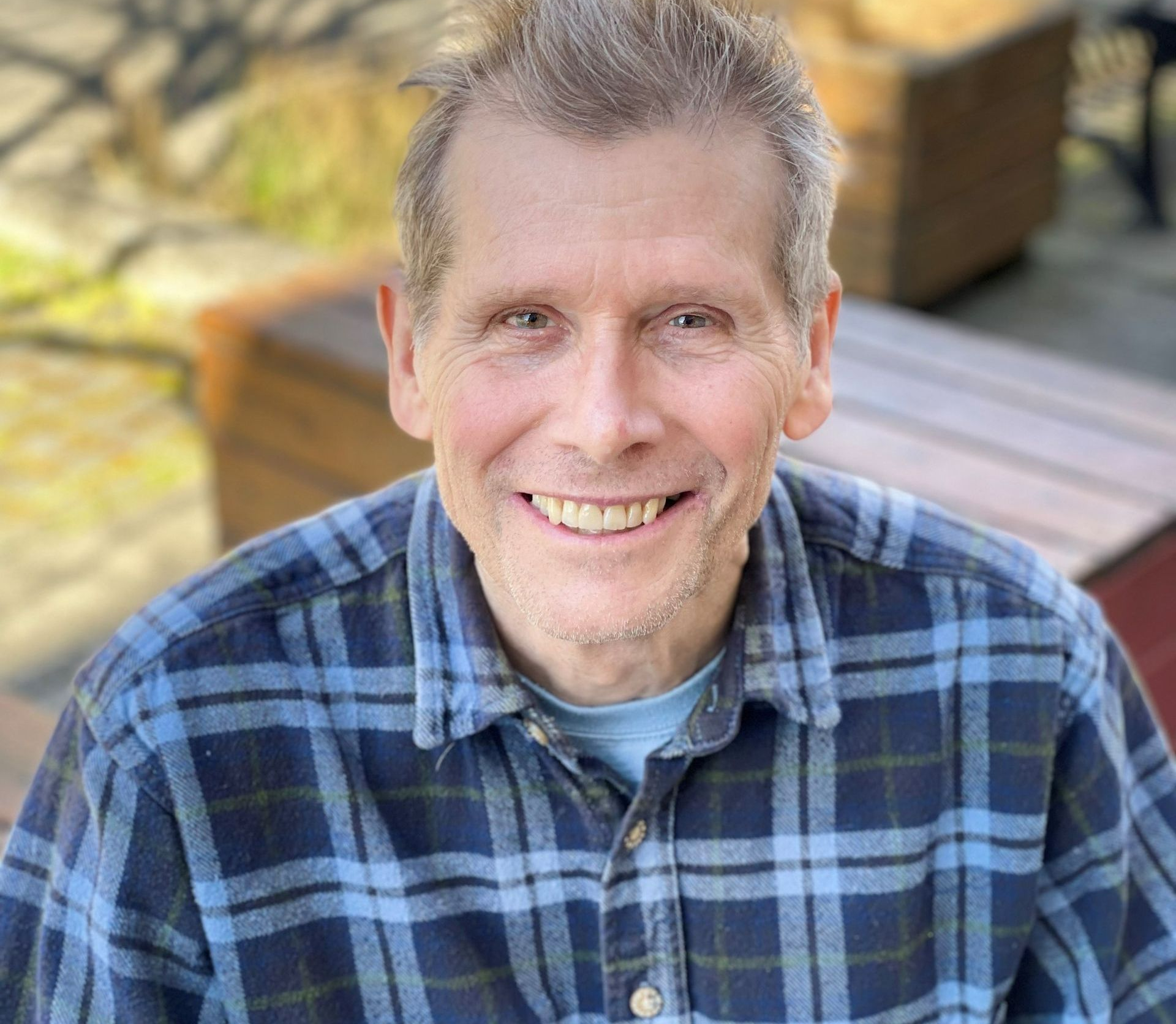 Middle-aged man with spiky gray hair smiles warmly, wearing a blue plaid shirt outdoors.