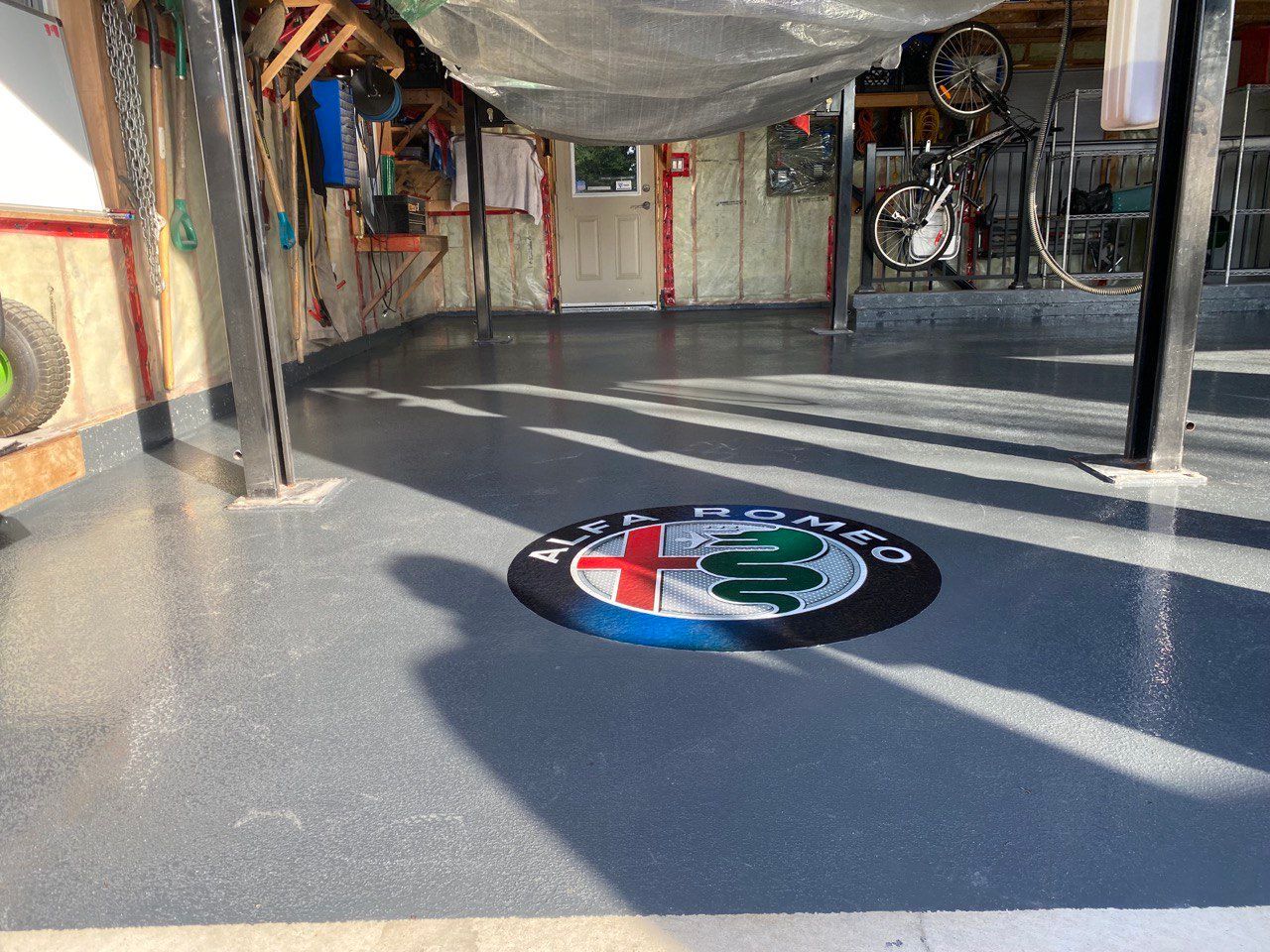 a garage that has epoxy flooring, coustom creation for the costumer with a branded logo of alfa romeo located at the center of the garage all coated with a layer of epoxy over it ensuring bright and shniy floors . the floors have a grey and blue epoxy tint that goes well with the cars in the garage. the logo consists of red, white, green and blue colours.