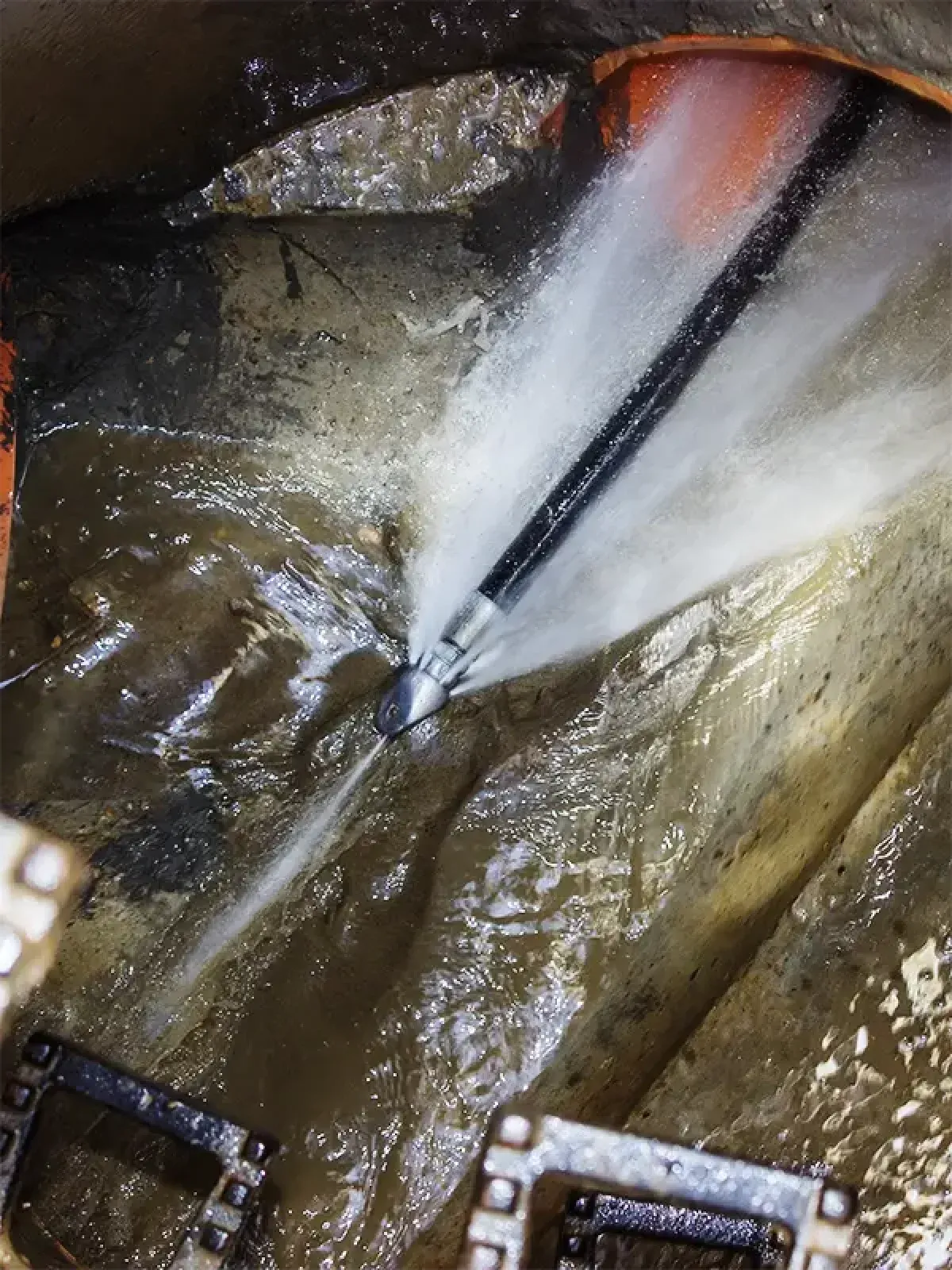 A high-pressure sewer jetter nozzle cleaning a pipe inside a manhole, spraying water forcefully against the walls.