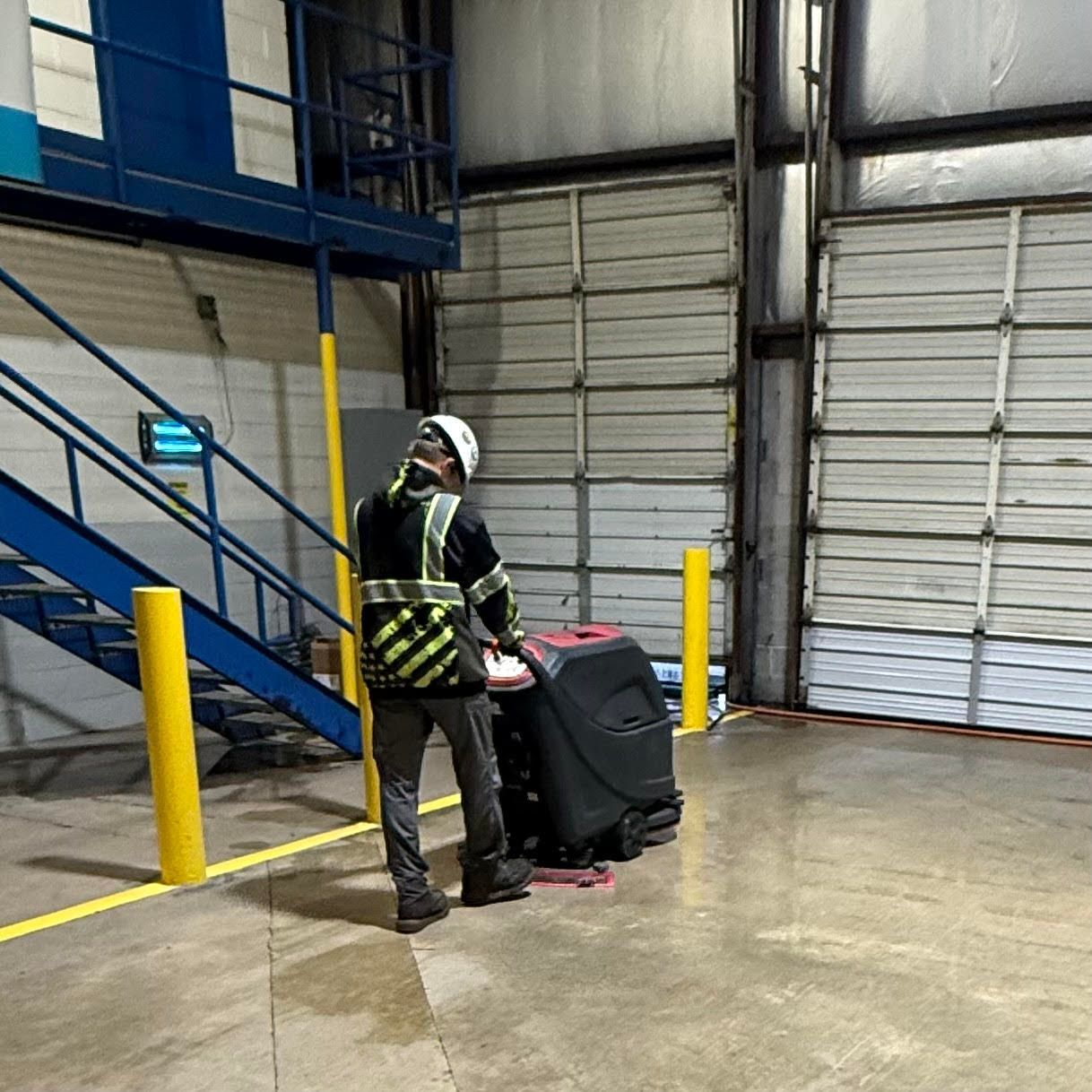 A person in a high-visibility vest and hard hat operates a floor-scrubbing machine in a warehouse facility.
