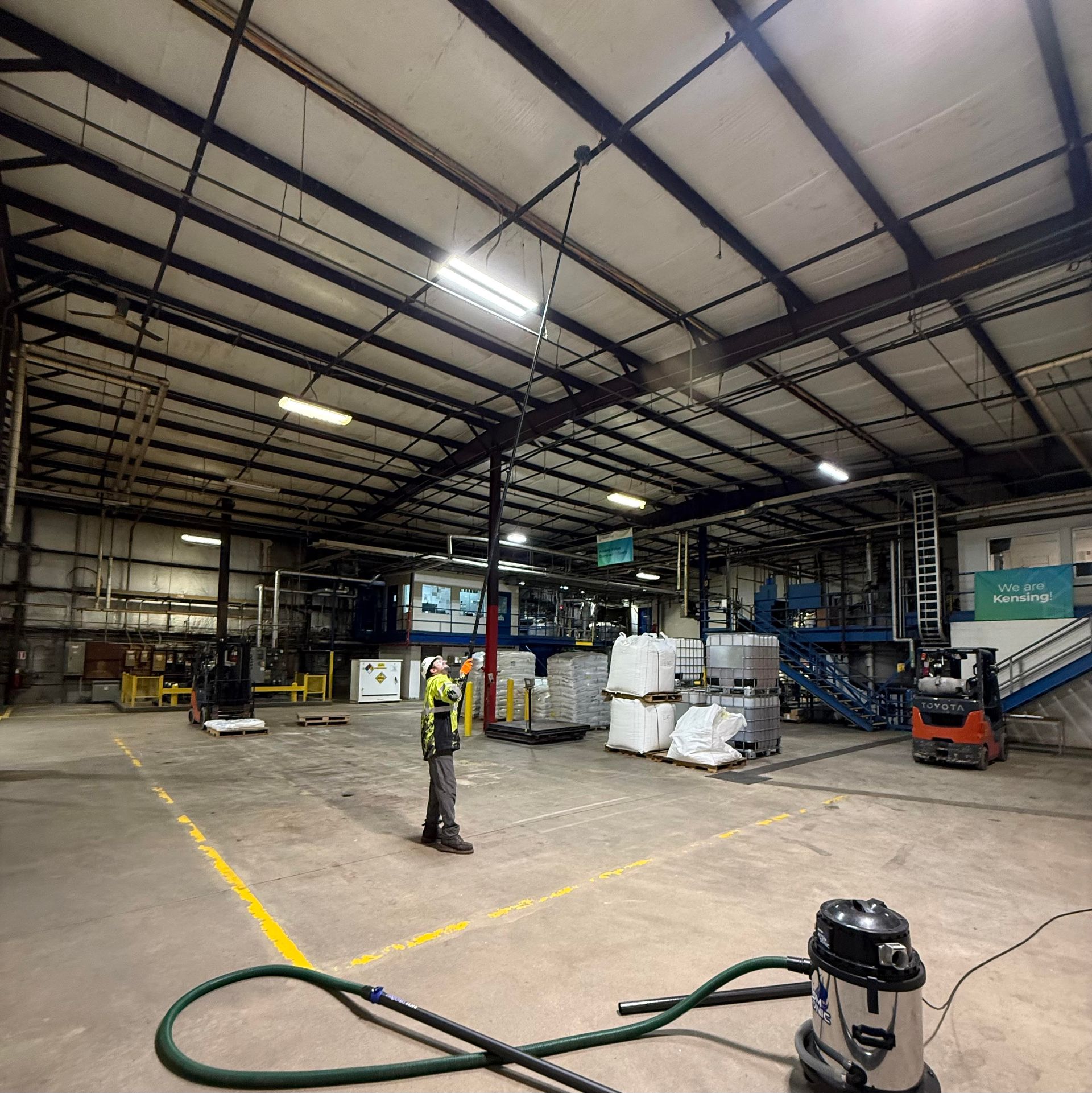 A person in a high-visibility vest stands in a large, industrial warehouse with stacked pallets and a shop vacuum nearby.
