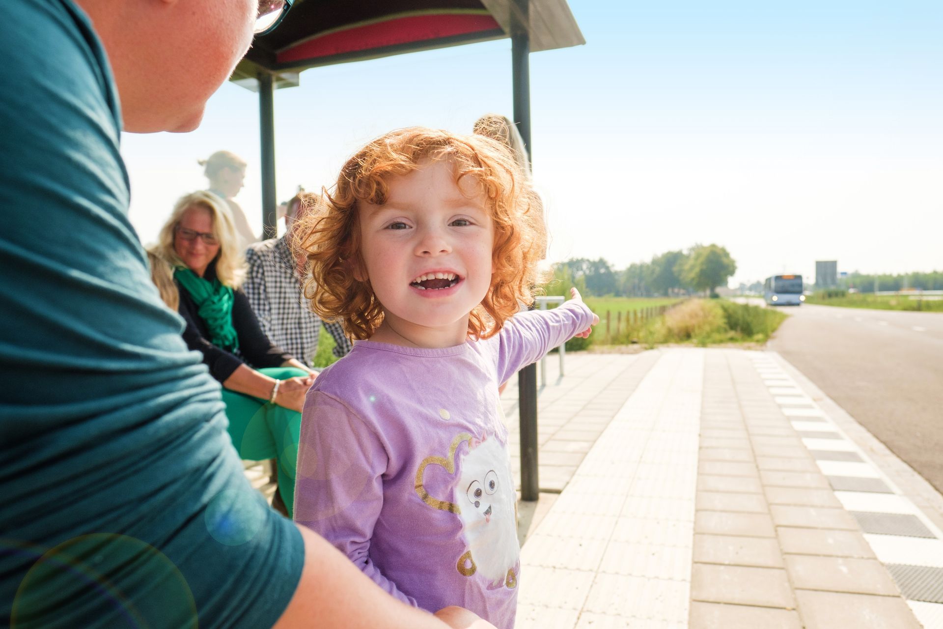 Vader met kind staan bij de halte. Het is zonnig. Het kind wijst naar de bus die aan komt rijden.