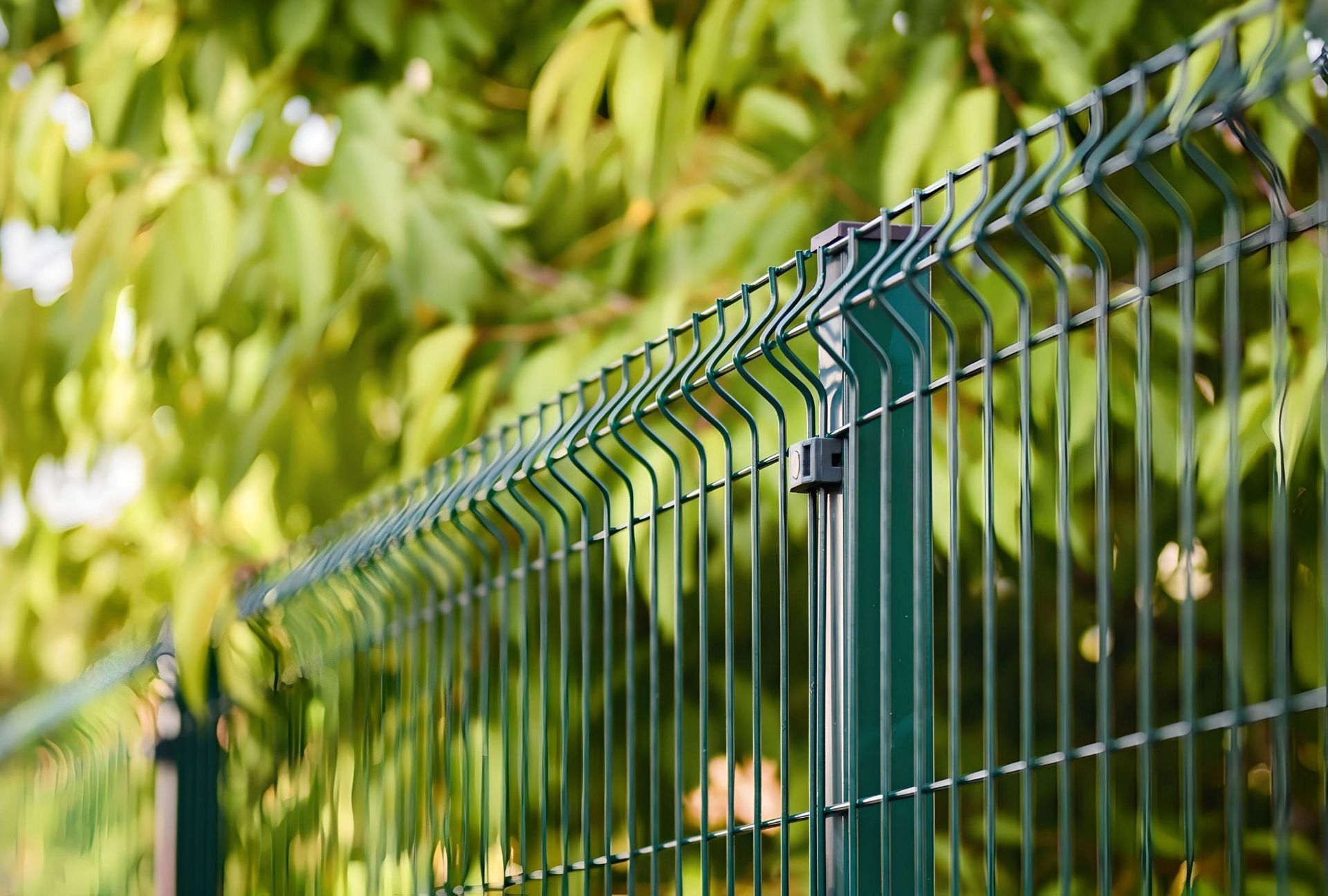 Green metal fence with diagonal wire mesh in front of blurred leafy foliage