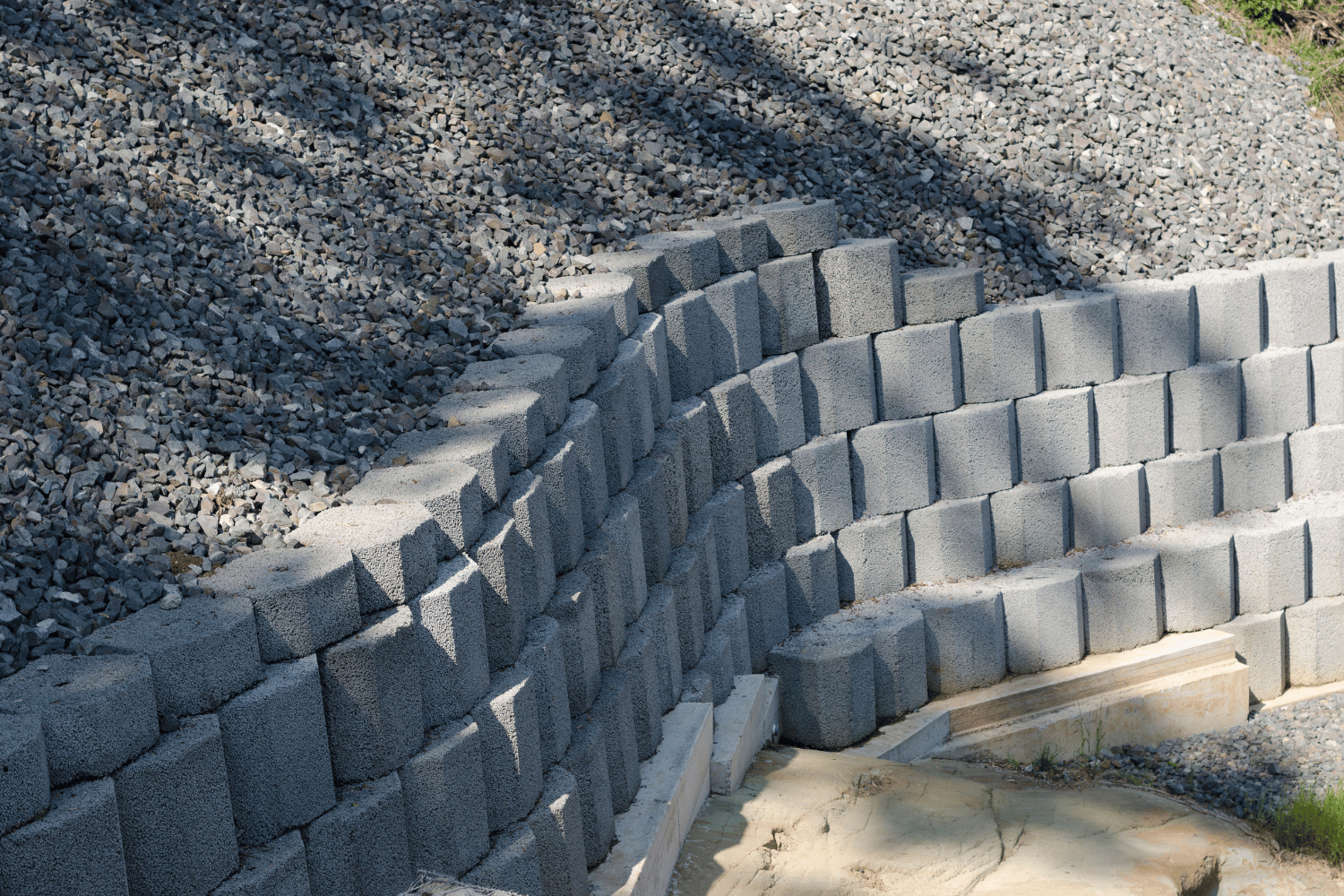 Curved gray retaining wall of interlocking blocks along a gravel slope beside sandy ground