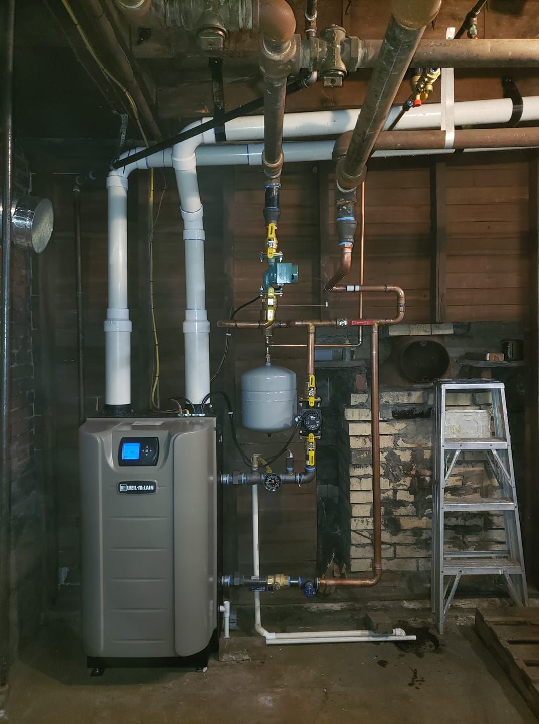 A boiler is sitting in a basement next to pipes and a ladder.