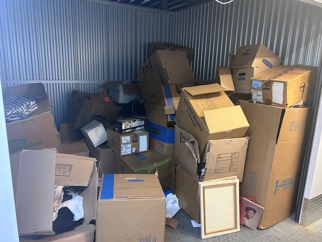 Storage unit filled with stacked cardboard boxes and household items in a corrugated metal room