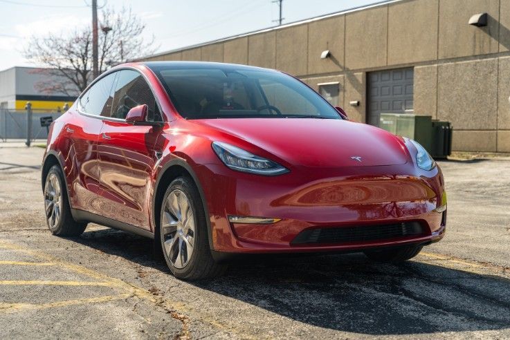 Red Tesla sedan parked on a sunlit street beside a beige industrial building