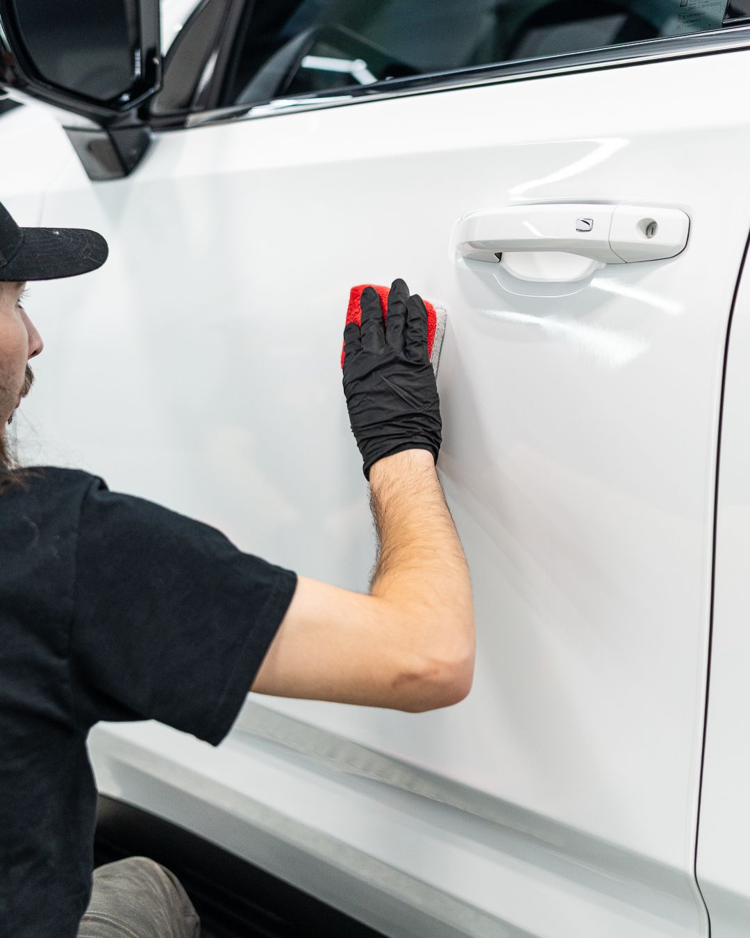 Person applying product to a white car door with a red applicator pad, wearing black gloves.