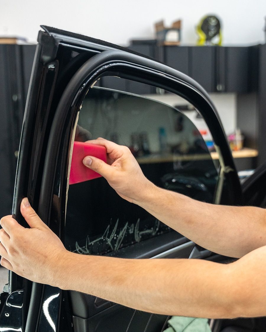 Hands use a pink squeegee to apply film on a car window in a garage.
