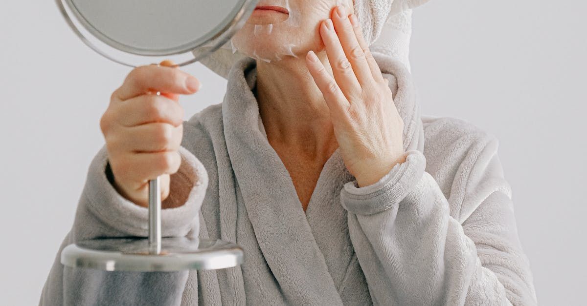 A woman in a bathrobe is looking at her face in a mirror.