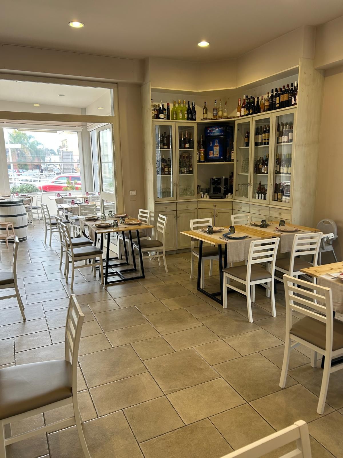 Interno del ristorante con tavoli e sedie bianchi, bottiglie di vino nelle vetrine, colori neutri.