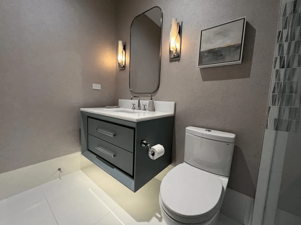 Modern bathroom with gray vanity, white toilet, mirror, wall sconces, and tiled shower corner