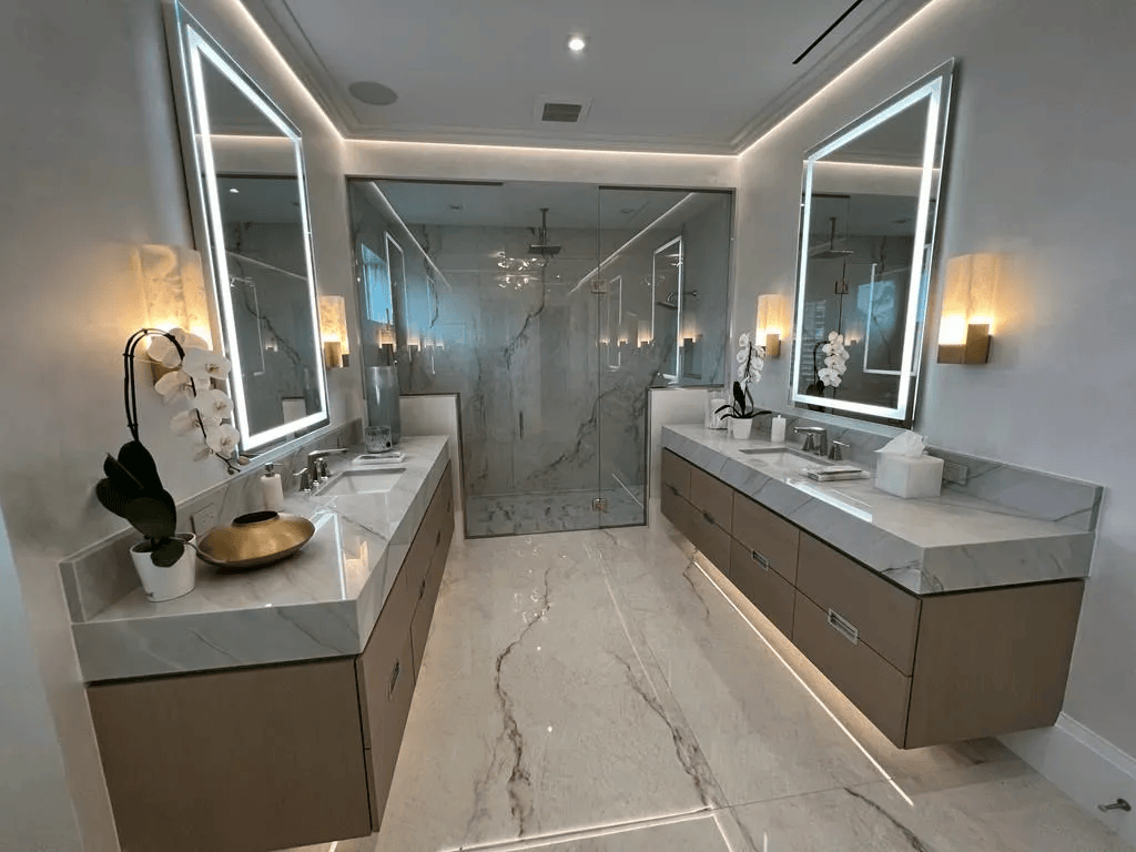 Modern bathroom with double sinks, illuminated mirrors, glass shower, and marble finishes.