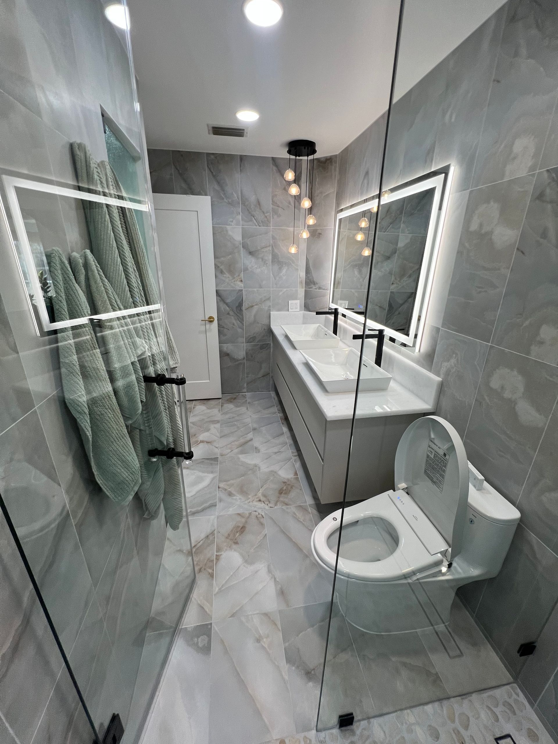 Narrow modern bathroom with glass shower, double sink vanity, and toilet on marble tile floors