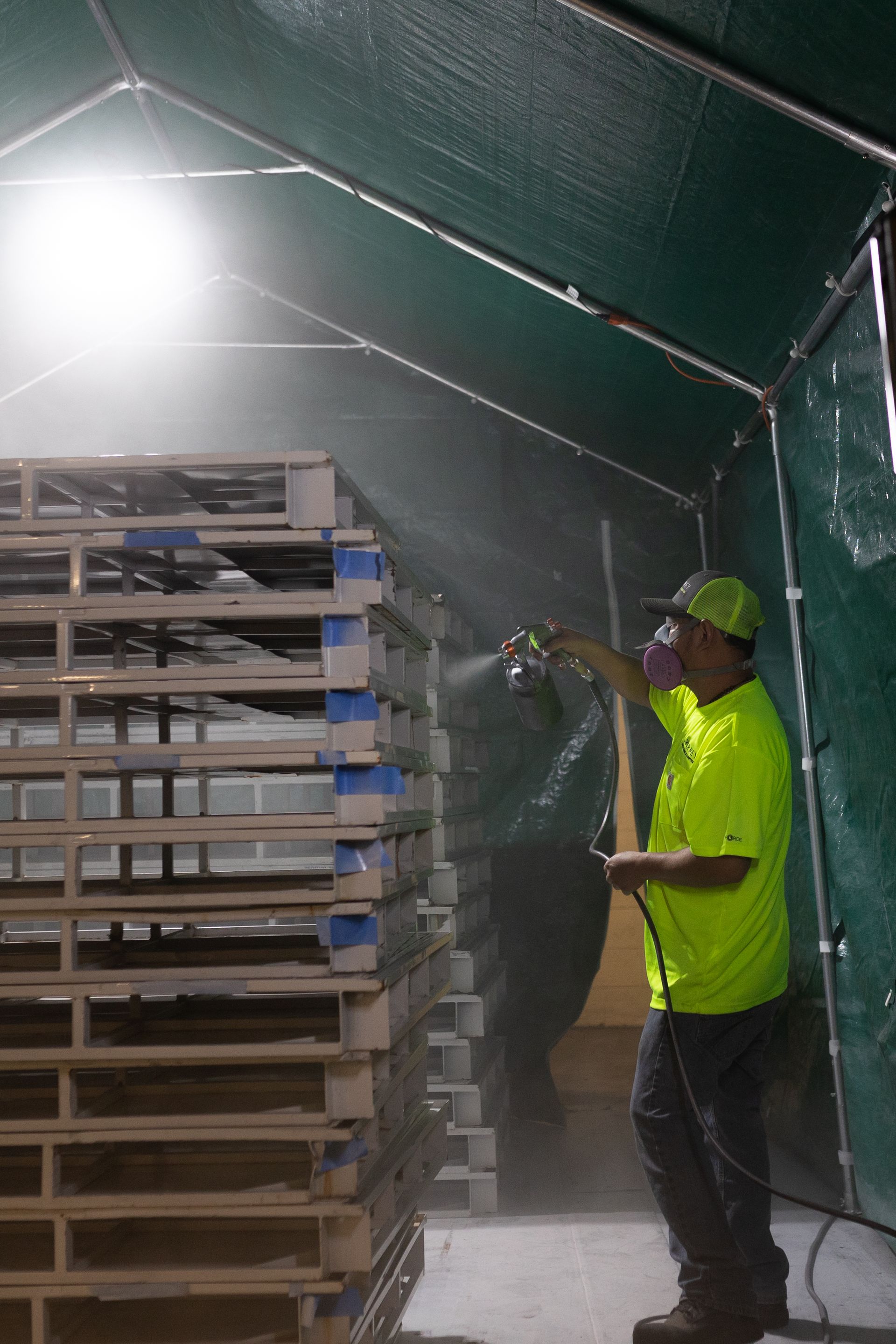 A man in a neon yellow shirt is spraying pallets in a tent.