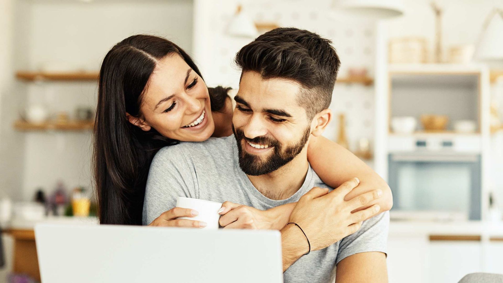 Smiling couple hugging in a bright kitchen, with the woman holding a mug.