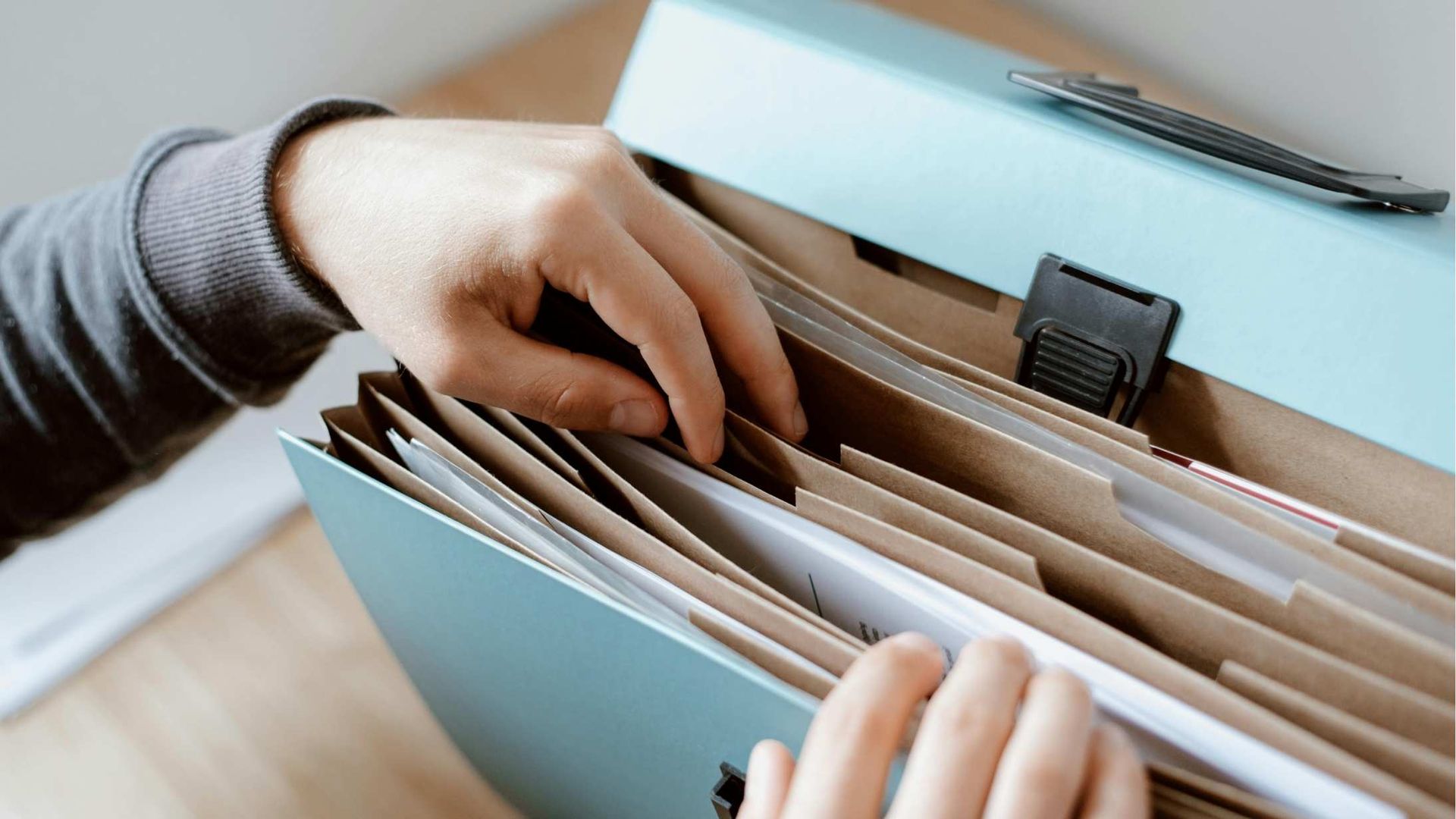 Hands sorting papers in a light blue file folder with a black clip on a desk.