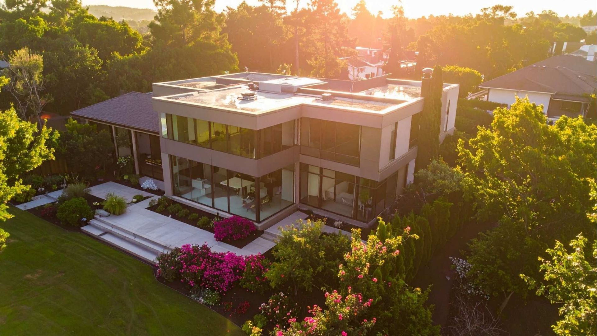 Sunlit modern two-story house surrounded by lush trees and pink flowers at sunset