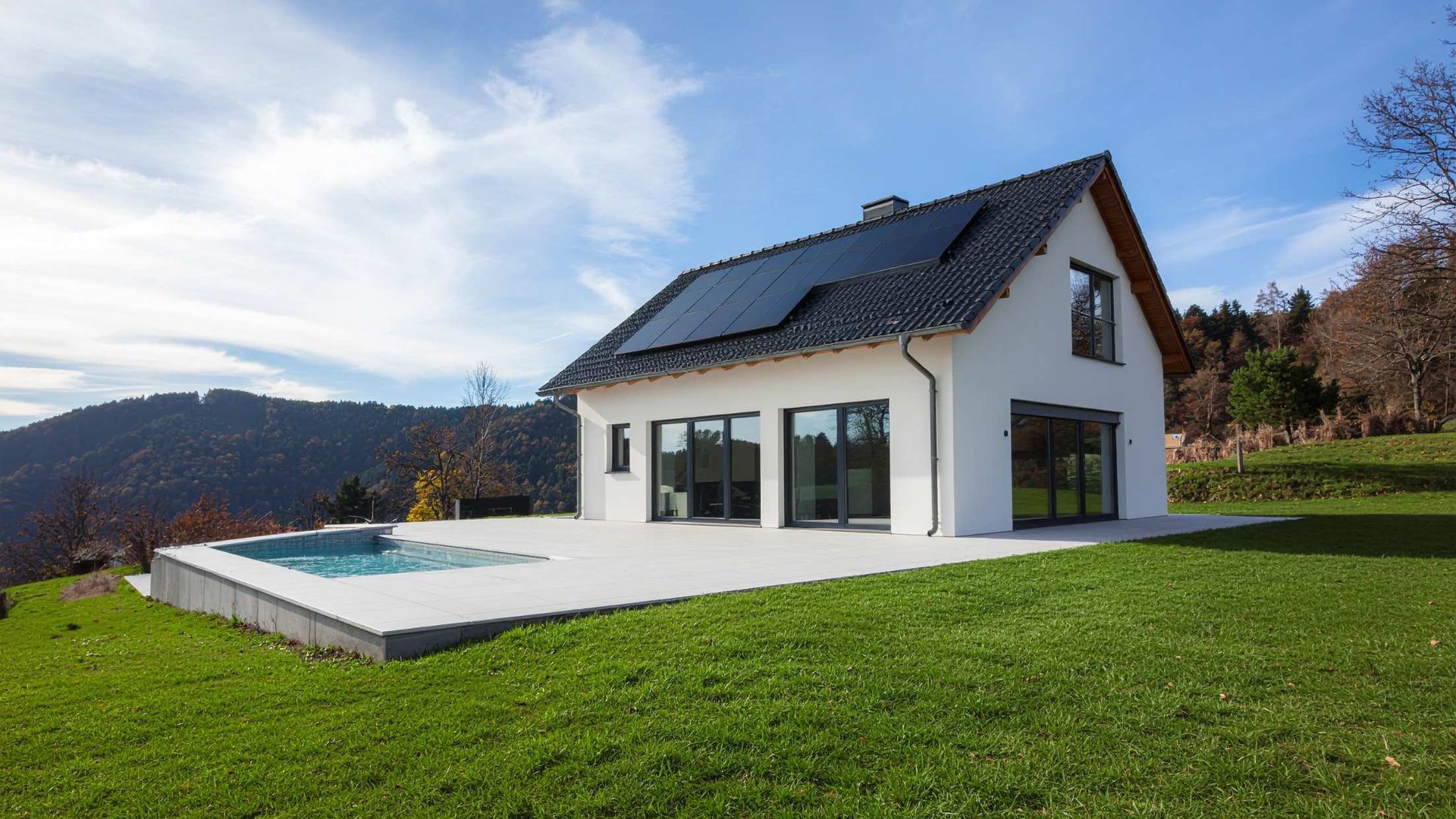 Modern white house with dark roof beside a backyard pool on a green lawn with mountains in the background