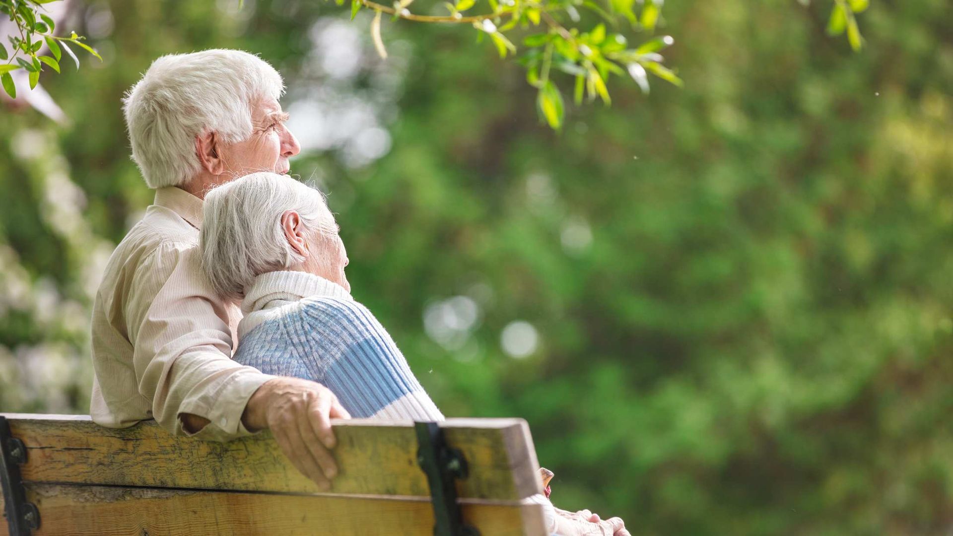 Two elderly people sit close together on a wooden bench in a green park, looking into the distance.