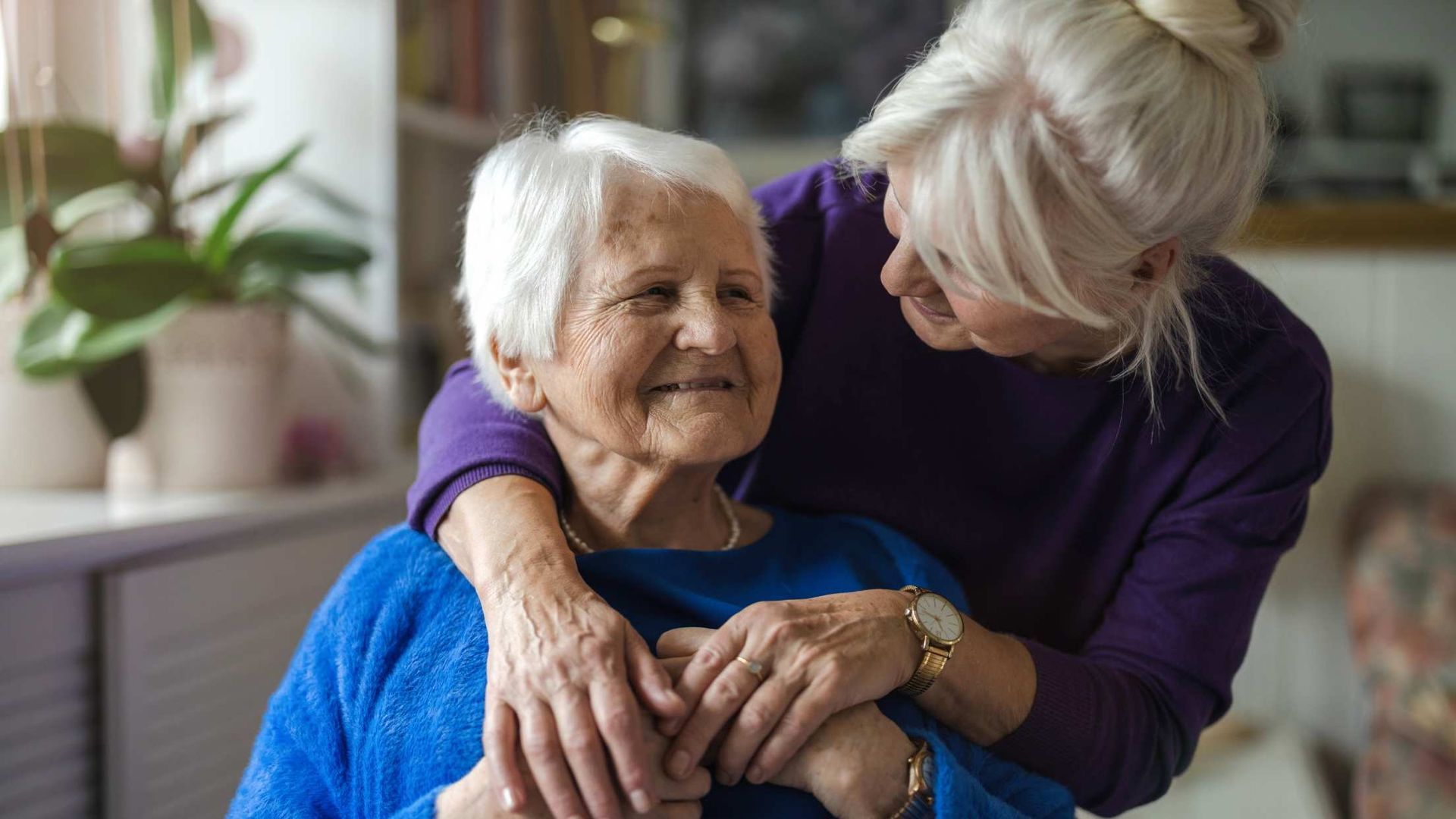 Older woman in a blue sweater embraces a smiling woman in purple indoors