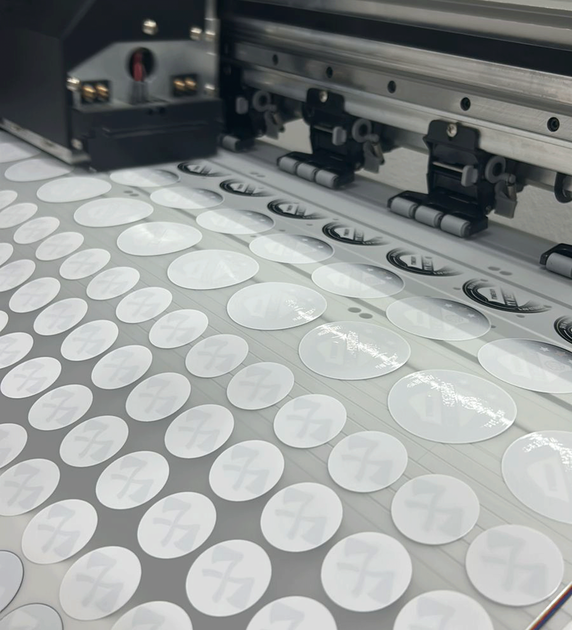 Uma impressora de grande formato recorta uma grade de adesivos circulares de uma folha de material vinílico branco.