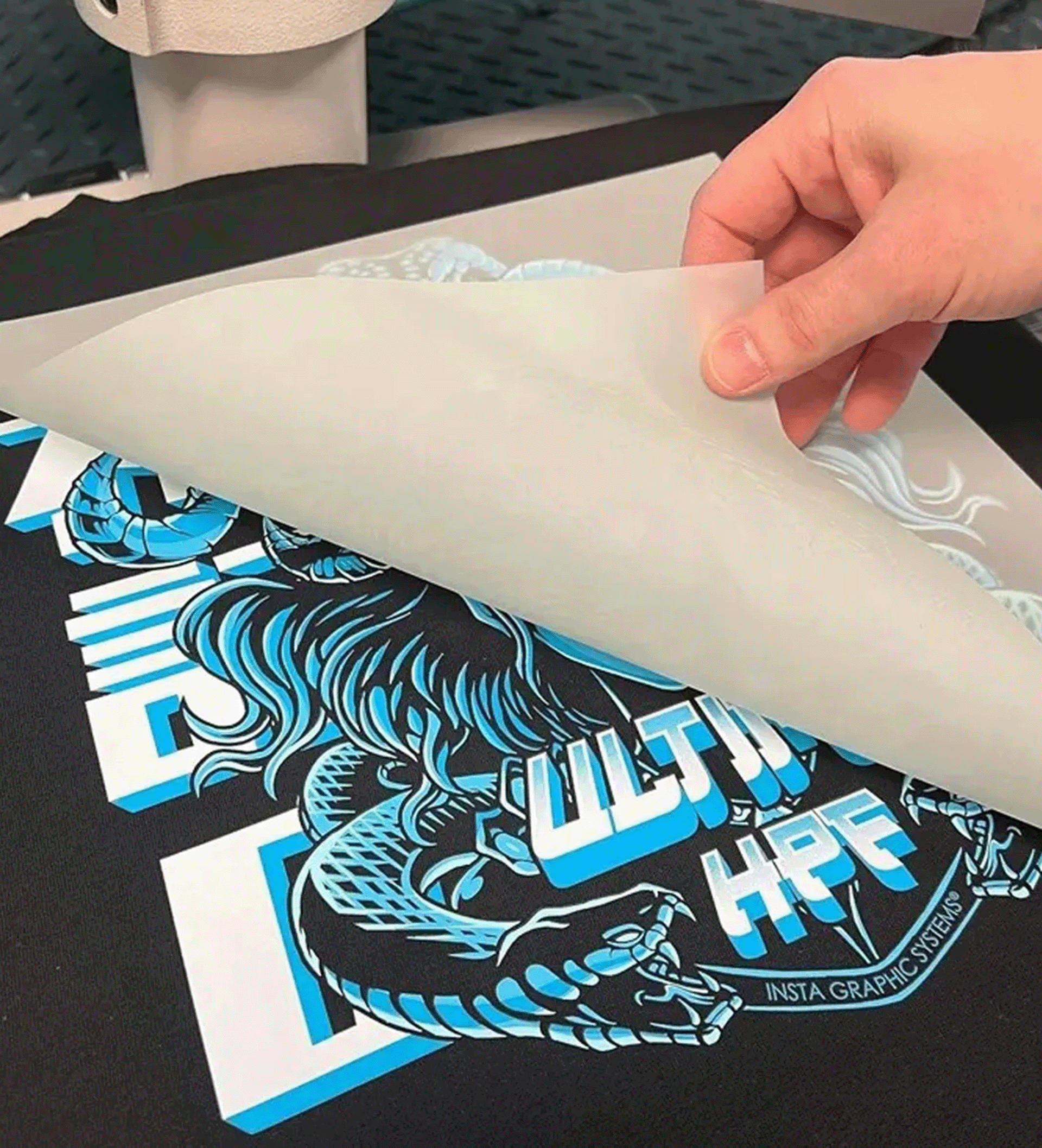 Uma mão retira uma folha de papel pergaminho de uma estampa serigráfica azul e branca em uma camiseta preta.