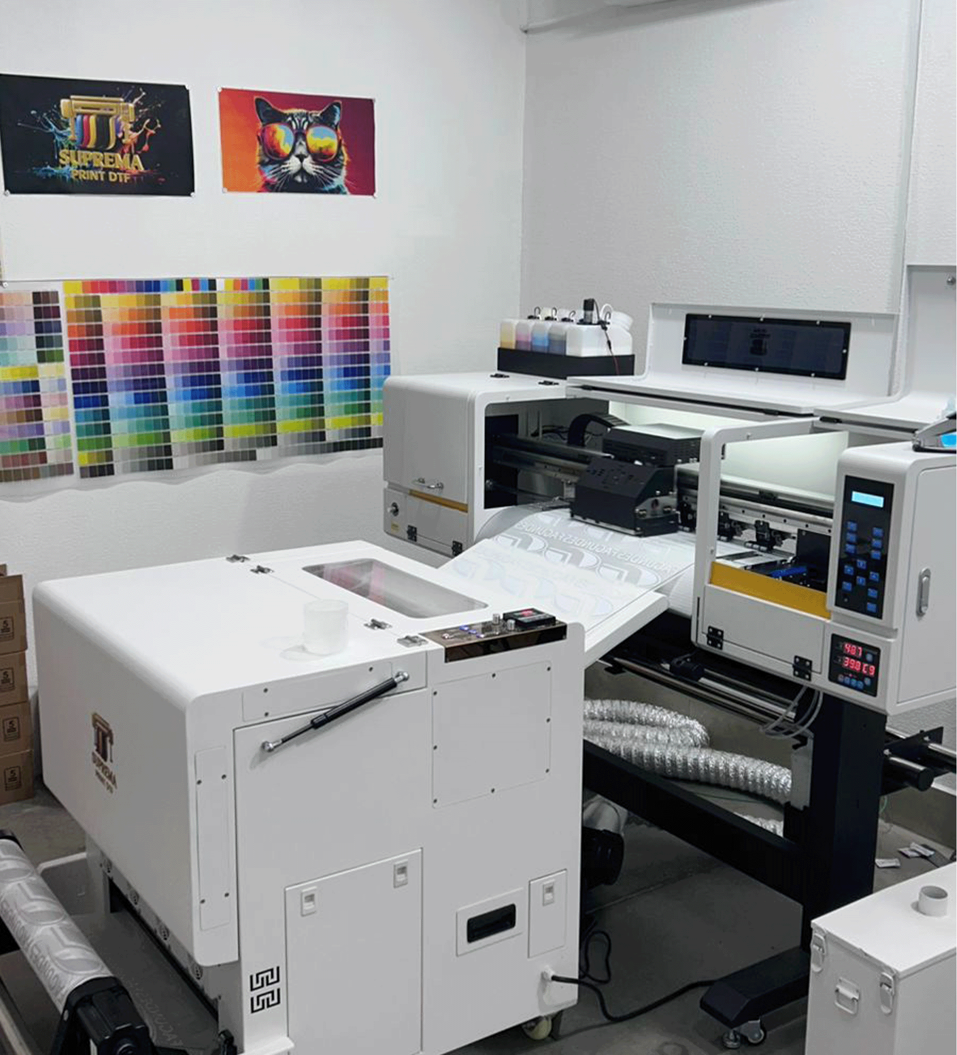 Uma grande impressora DTF industrial em uma oficina, com um painel de cores na parede e impressões de teste acima.
